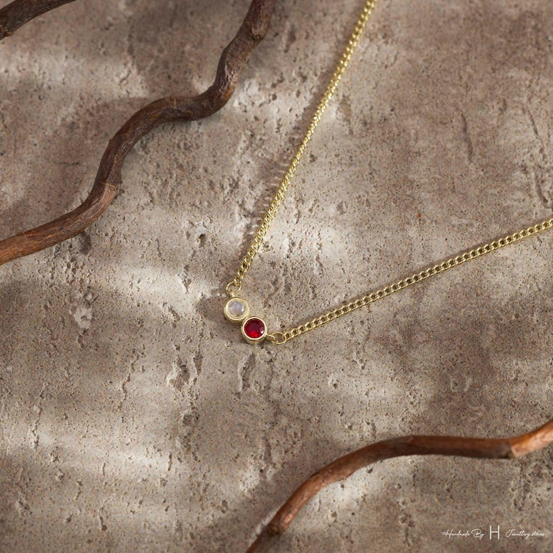 May include: A gold-tone chain necklace featuring two small, circular bezel-set stones. One stone is a translucent white gem, and the other is a vibrant red gem. The necklace is displayed with a thin, curved brown branch.