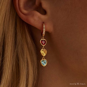 May include: Gold hoop earrings with a dangling charm featuring four heart-shaped gemstones in red, yellow, and blue.