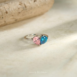 May include: A silver-toned ring featuring two pear-cut gemstones arranged to form a heart shape. One gemstone is light pink and the other is a vibrant blue. The band is thin and metallic, showcasing a polished finish that reflects light.