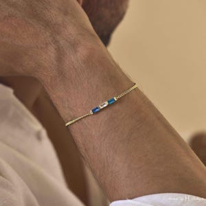 May include: A delicate gold chain bracelet featuring three rectangular baguette-cut stones arranged horizontally in the center. The stones consist of two deep blue gems on the outer sides and one clear, colorless gem in the middle, all set in gold.