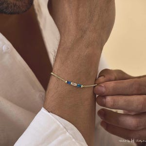 May include: A delicate gold chain bracelet featuring a central horizontal arrangement of three rectangular gemstones. The center stone is clear, flanked by two vibrant blue stones, all set in a thin, polished gold metal frame on a fine link chain.
