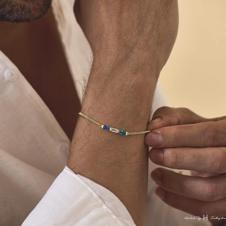 May include: A delicate gold chain bracelet featuring a central horizontal arrangement of three rectangular gemstones. The center stone is clear, flanked by two vibrant blue stones, all set in a thin, polished gold metal frame on a fine link chain.