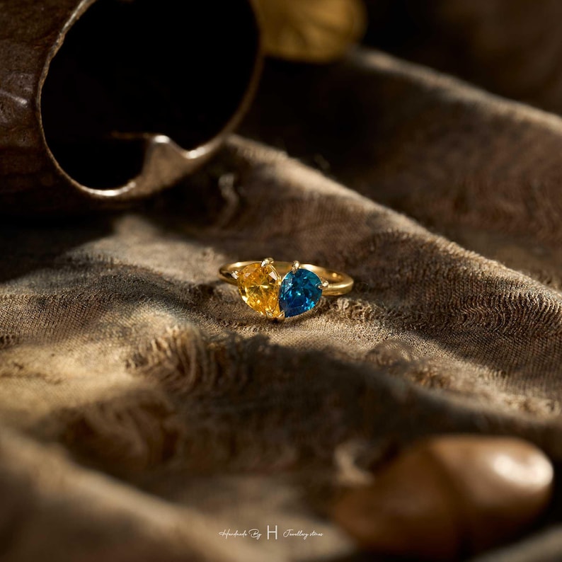 May include: A gold ring featuring two heart-shaped gemstones, one yellow and one blue, set side-by-side. The band is thin and metallic gold. The text 'Handmade by H Jewellery store' is printed in small white letters at the bottom of the image.