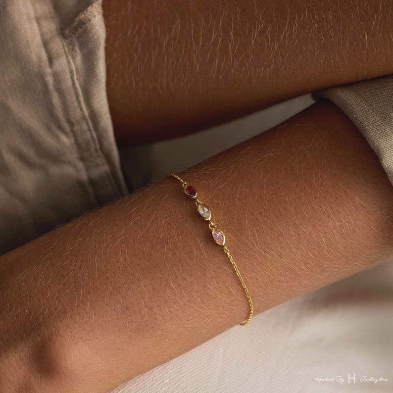 May include: A delicate gold chain bracelet featuring three marquise-cut gemstones in a row. The stones are set in gold bezels and include a deep red, a clear white, and a soft pink gem. The bracelet has a fine link chain design.