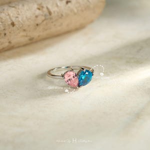 May include: A silver-toned ring featuring two pear-shaped gemstones, one pink and one blue, set side-by-side. The text Oct stone pink is positioned near the pink gem, and Dec stone is near the blue gem. The band is thin and metallic.