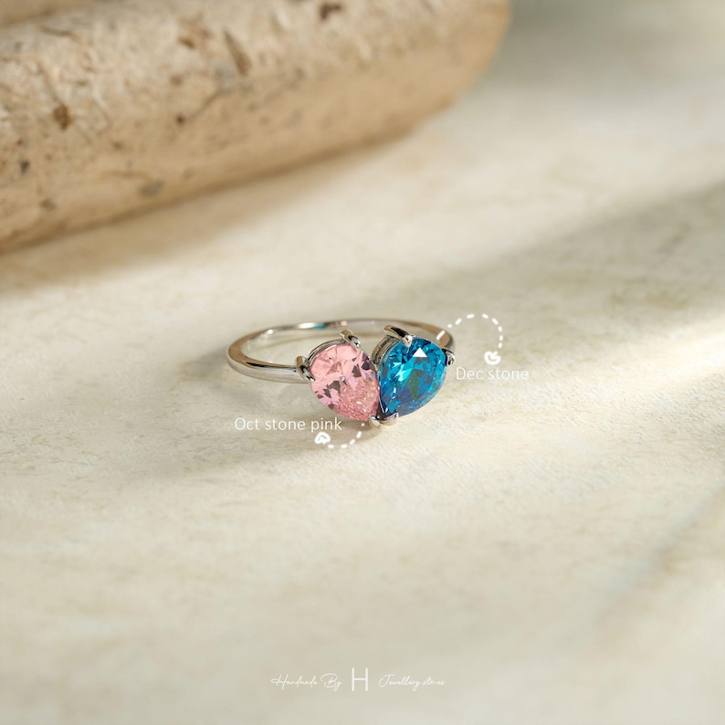 May include: A silver-toned ring featuring two pear-shaped gemstones, one pink and one blue, set side-by-side. The text Oct stone pink is positioned near the pink gem, and Dec stone is near the blue gem. The band is thin and metallic.