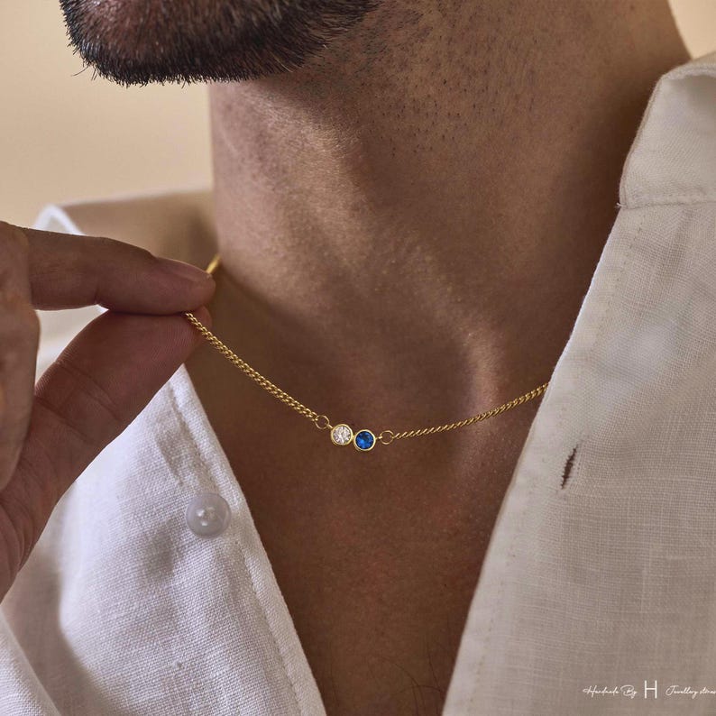 May include: A gold-toned chain necklace featuring two small, circular bezel-set stones. One stone is clear and white, while the other is a vibrant blue. The necklace rests against a white collared shirt, showcasing a delicate and refined design.
