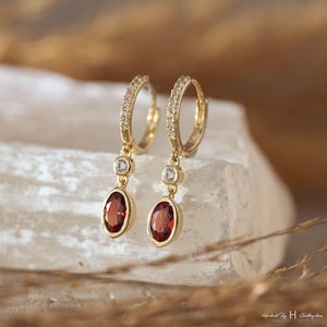 Peut inclure: Une paire de boucles d'oreilles créoles dorées avec des pierres précieuses ovales brun-rouge. Les créoles sont incrustées de petites pierres claires. Chaque boucle d'oreille a une petite pierre claire ronde au-dessus de la pierre ovale.