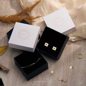 May include: Two square white gift boxes with sunburst logos, one open to reveal gold stud earrings with rectangular green stones, and another open to show a delicate gold chain bracelet with small colorful stones, alongside gold necklaces and rings.