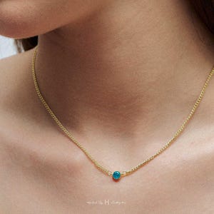 May include: A delicate gold-tone chain necklace featuring a single, small, round, vibrant turquoise-blue stone pendant centered on the chain. The necklace rests against the skin, showcasing a simple link design with a polished metal finish.
