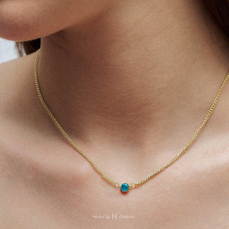 May include: A delicate gold-tone chain necklace featuring a single, small, round, vibrant turquoise-blue stone pendant centered on the chain. The necklace rests against the skin, showcasing a simple link design with a polished metal finish.