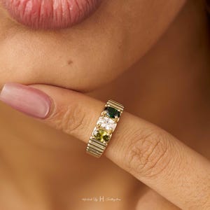 May include: A gold-toned ring featuring three oval-cut stones arranged horizontally. The centre stone is clear and colourless, flanked by a dark green stone on one side and a light olive green stone on the other. The band has a ribbed, textured finish.