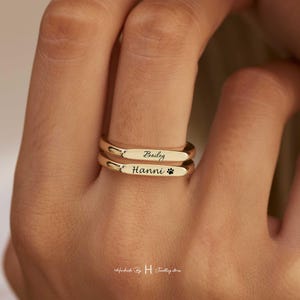 Op de afbeelding: Een set van twee gestapelde goudkleurige ringen met een gladde, gepolijste afwerking. De bovenste ring is gegraveerd met de naam Bailey in cursief schrift en de onderste ring met de naam Hanni, gevolgd door een klein pootafdruk-icoon.