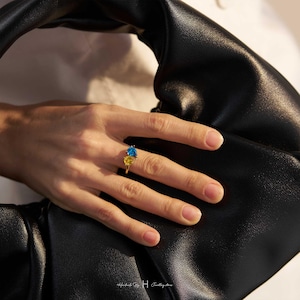 May include: A gold ring featuring two faceted gemstones, one pear-shaped blue stone and one round yellow stone, set on a thin gold band. The ring is positioned against a black leather accessory. The text Handmade By H Jewellery Store is visible.