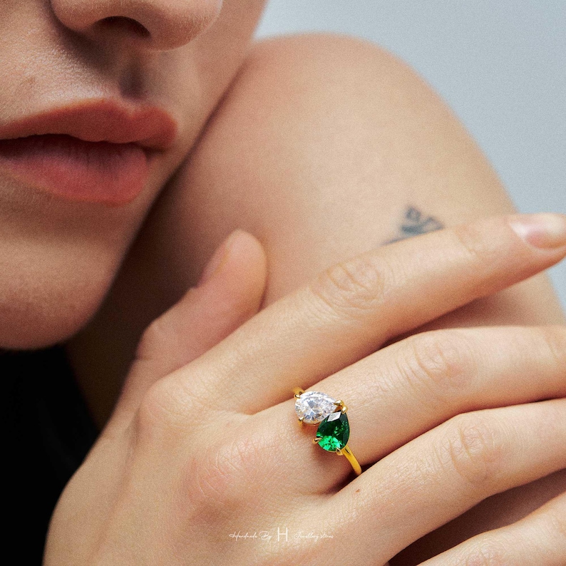 May include: A gold ring featuring two pear-cut stones set side-by-side. One stone is clear and colorless, while the other is a vibrant emerald green. The band is thin and polished gold, showcasing a minimalist design that highlights the contrasting gems.