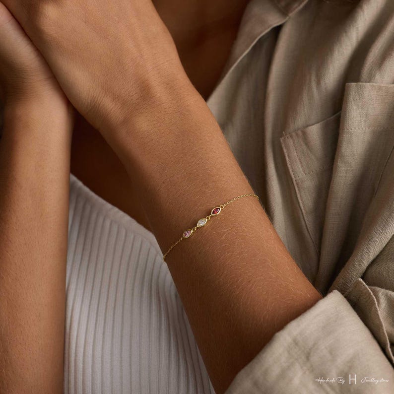 May include: A delicate gold chain bracelet featuring three marquise-cut gemstones in a row. The stones include a light pink gem, a clear white gem, and a deep pink gem, each set in a gold bezel. The bracelet has a thin, polished gold link chain.