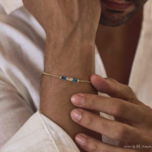 May include: A delicate gold chain bracelet featuring three rectangular baguette-cut stones arranged horizontally in the center. The stones consist of a clear white center stone flanked by two vibrant blue stones, all set in a thin, polished gold-tone metal chain.