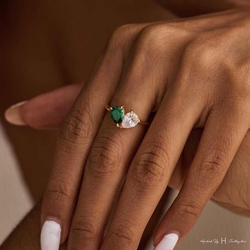 May include: A gold ring featuring two pear-cut stones set side-by-side on a thin band. One stone is a deep green, and the other is a clear, colorless stone. The ring is positioned on a finger, showcasing the dual-stone design and metal finish.