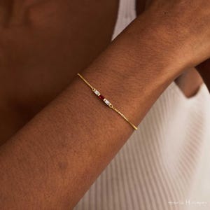 Può includere: Un delicato bracciale a catena dorata caratterizzato da una disposizione orizzontale centrale di tre pietre taglio baguette. La pietra centrale è di un rosso vivace, affiancata da due pietre chiare e incolori, incastonate in oro.
