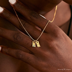 Puede incluir: Collar de oro con dos pequeños colgantes rectangulares. Cada colgante tiene una letra, "M" y "K", y una pequeña piedra preciosa.