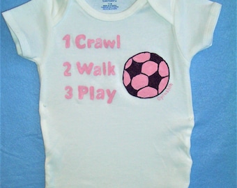 May include: White baby onesie with pink text that reads "1 Crawl 2 Walk 3 Play" and a pink and black soccer ball graphic.