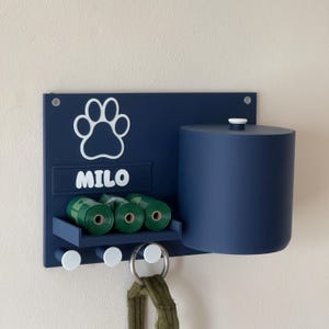 Peut inclure: Un distributeur de sacs à déjections canines mural bleu avec une empreinte de patte et le nom "MILO" dessus. La station comprend un distributeur de sacs à déjections canines, un crochet pour laisse et trois crochets pour d'autres articles.