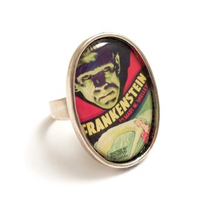 May include: A silver ring with a round, glass-like oval cameo featuring a vintage movie poster for Frankenstein. The poster features a black and white image of the monster with the title "Frankenstein by Mary W. Shelley" in red and yellow letters.