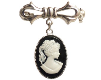 Op de afbeelding: Zilverkleurige strikbroche met een hangende ovale camee. De camee toont een wit profiel op een zwarte achtergrond. Deze broche is een klassiek accessoire, perfect om een vleugje vintage elegantie aan elke outfit toe te voegen.