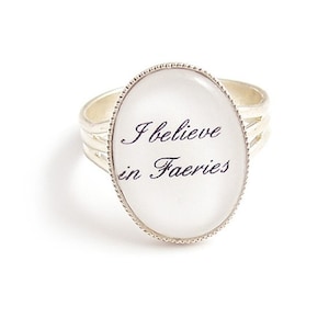 May include: A silver ring with a white oval-shaped stone that says "I believe in Faeries" in black lettering.
