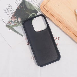 May include: A black phone case with a camera cutout, resting on a white surface with printed text and a wooden cutting board. The case is smooth and appears to be made of a durable material.