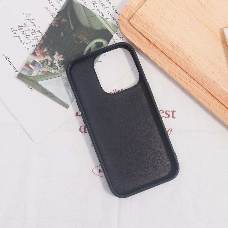 May include: A black phone case with a camera cutout, resting on a white surface with printed text and a wooden cutting board. The case is smooth and appears to be made of a durable material.