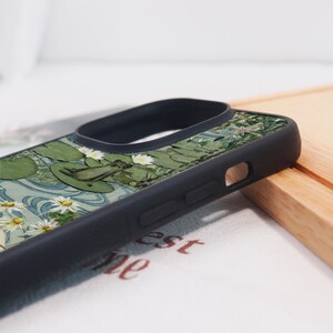 May include: Black phone case featuring a detailed illustration of a frog on a lily pad, surrounded by water lilies and daisies. The design has a vintage, artistic style with green, white, and blue hues. The case has a black border and a raised camera ring.
