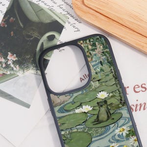 May include: A phone case featuring a whimsical design with a frog sitting on a lily pad, surrounded by water lilies and daisies. The case has a dark gray border and a cutout for the camera.
