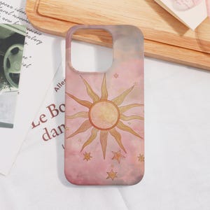 May include: A phone case with a celestial design. The case has a pink and peach background, with a sun and star graphic. The sun is in the centre, with rays extending outwards. Several stars are scattered around the sun.