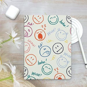 May include: A cream-colored tablet case covered in colorful smiley face emojis and exclamations like "Wow!" and "Hehe!". The case has a white border and is surrounded by a white pen, a white charging case, and a daisy.