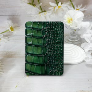 May include: A rectangular card holder featuring a deep forest green crocodile skin texture. The surface displays a gradient of dark to light green tones with a glossy finish, showcasing a realistic embossed pattern of large and small scales.