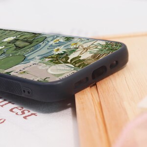 May include: A black phone case featuring a detailed illustration of a pond with lily pads, white flowers, and a frog. The design has a vintage, artistic style with the word "healing" in script.
