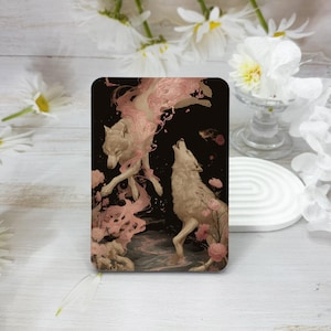 May include: A rectangular art print depicting two white wolves on a black background. One wolf leaps, the other howls. Pink floral details and flowing pink elements enhance the artwork's design.