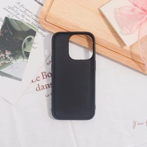 May include: A black phone case with a camera cutout. The case is made of a smooth material and is designed to protect the phone. The case is on a white surface with a wooden cutting board and a card with a pink ribbon in the background.