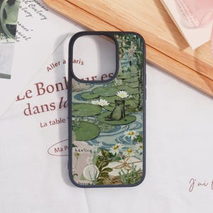 May include: A phone case featuring a serene pond scene with a frog on a lily pad, white flowers, and a blue water background. The case has a black border and includes the text "healing".
