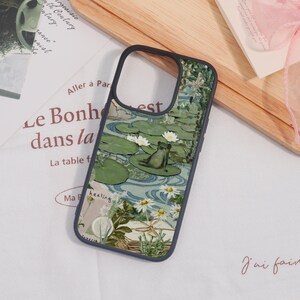 May include: A phone case with a nature-themed design. The case features a frog sitting on a lily pad in a pond, surrounded by water lilies and other aquatic plants. The case has a black border.