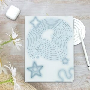 May include: A white notebook with a blue abstract design of wavy lines and stars. A white stylus and a white wireless charging pad are next to the notebook. Daisies and a white earbud case are also in the image.