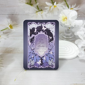 Könnte beinhalten: Ein lila und schwarzer Buchumschlag mit einem verzierten Design, das einen schwarzen Wolf und Schmetterlinge zeigt. Der Titel des Buches lautet "One Dark Window" mit dem Untertitel "A paranormal mystery" und dem Namen des Autors "Rachel Gillig".
