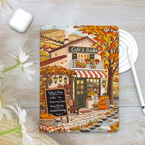 May include: A rectangular notebook featuring a colorful illustration of a cozy cafe and bookstore with autumn foliage, a menu board listing coffee, tea, pastries, cakes, soup, and books, a balcony with potted flowers, and a small stream in the foreground.