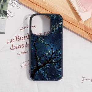 May include: A black phone case featuring a dark blue night scene with a tree and white and blue flowers. The case has a black border and is on a white surface with French text.