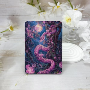 May include: A rectangular art print featuring a pink snake coiled around a tree in a moonlit forest. The artwork has a fantasy theme with pink flowers and a full moon in the background. The color palette includes shades of pink, purple, and blue.