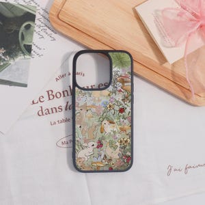 May include: A phone case with a whimsical forest scene featuring rabbits, mushrooms, and butterflies in soft pastel colors. The case has a black border and a cutout for the camera. The design includes the text "Aller à aris".