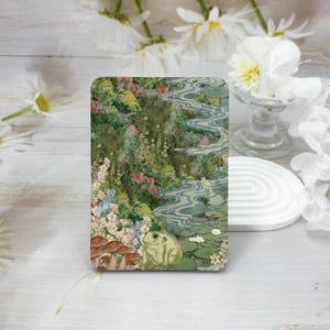 May include: A green frog sits on a bed of lily pads in a whimsical illustration. The frog is surrounded by a blue stream, green foliage, and mushrooms.