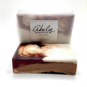 May include: Artisan soap bar with a layered design in shades of brown and white. The soap is rectangular and has a marbled appearance. The label reads "Abela ARTISAN NEW YORK" and indicates a weight of 6.0 oz / 141g.