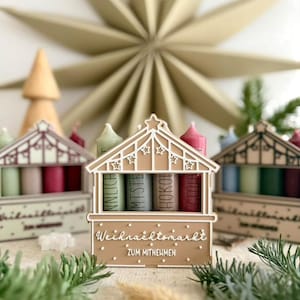 May include: A festive Christmas market-themed display featuring miniature wooden stalls with colorful candles. The stalls are decorated with intricate cut-out designs and the text "Weihnachtsmarkt ZUM MITNEHMEN". A large star-shaped ornament is in the background.
